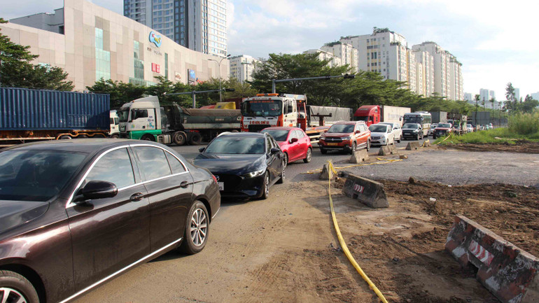 Phía bên trái là công trường, phía phải là cảnh xe tải, ô-tô, container xếp hàng dài di chuyển qua khu vực này.