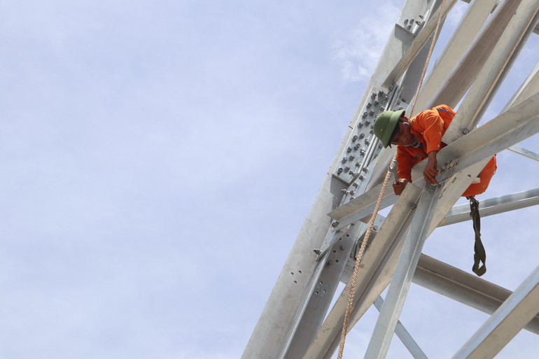 Công nhân đu mình trên cột kim loại nóng, cẩn thận kiểm tra, siết chặt từng bu-lông.