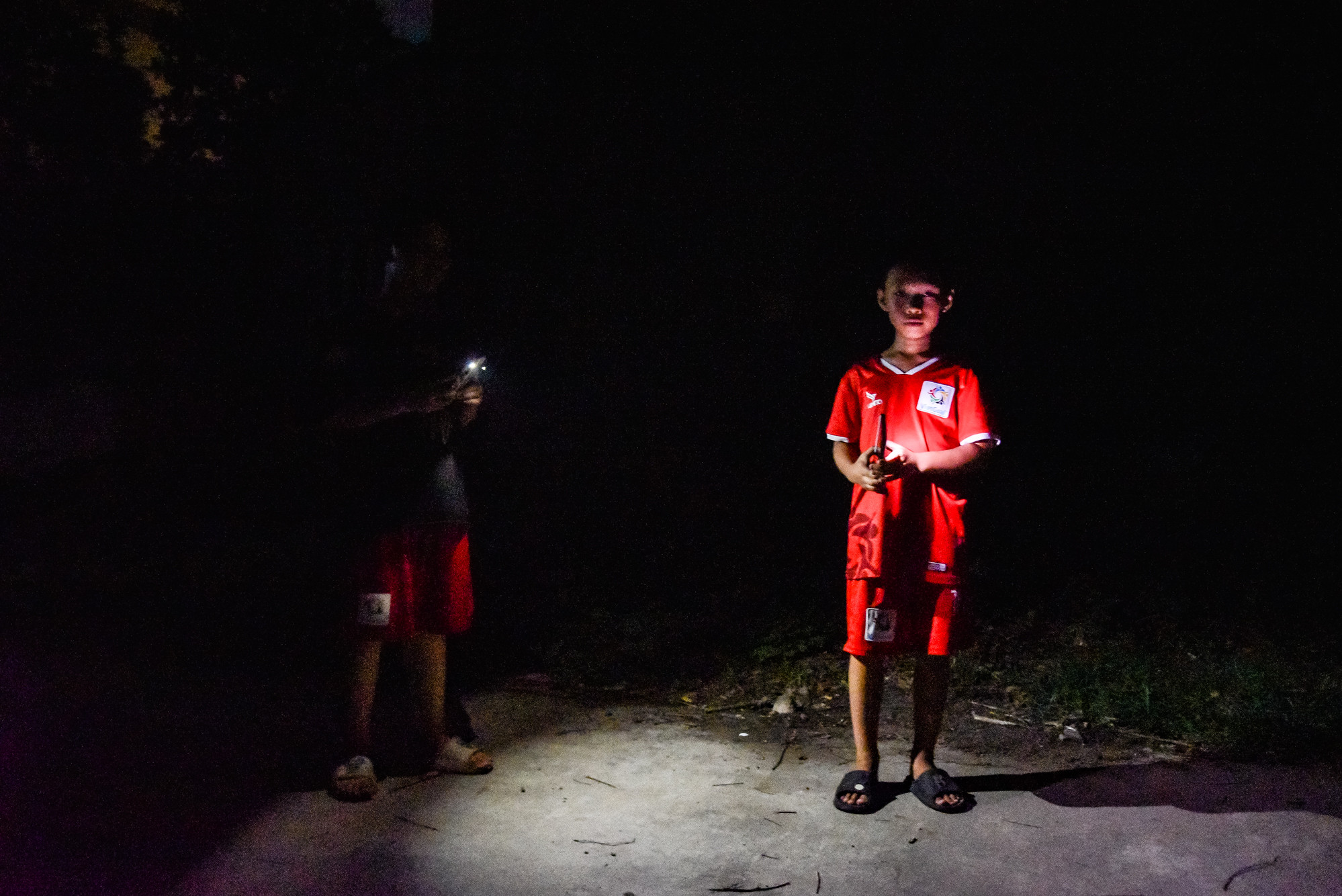 Nhiều khu vực trong thôn tối đen, không có đèn đường, hai bạn nhỏ đã phải sử dụng đèn pin của điện thoại để soi sáng đường đi. Nhiều khu vực trong thôn tối đen, không có đèn đường, hai bạn nhỏ đã phải sử dụng đèn pin của điện thoại để soi sáng đường đi.