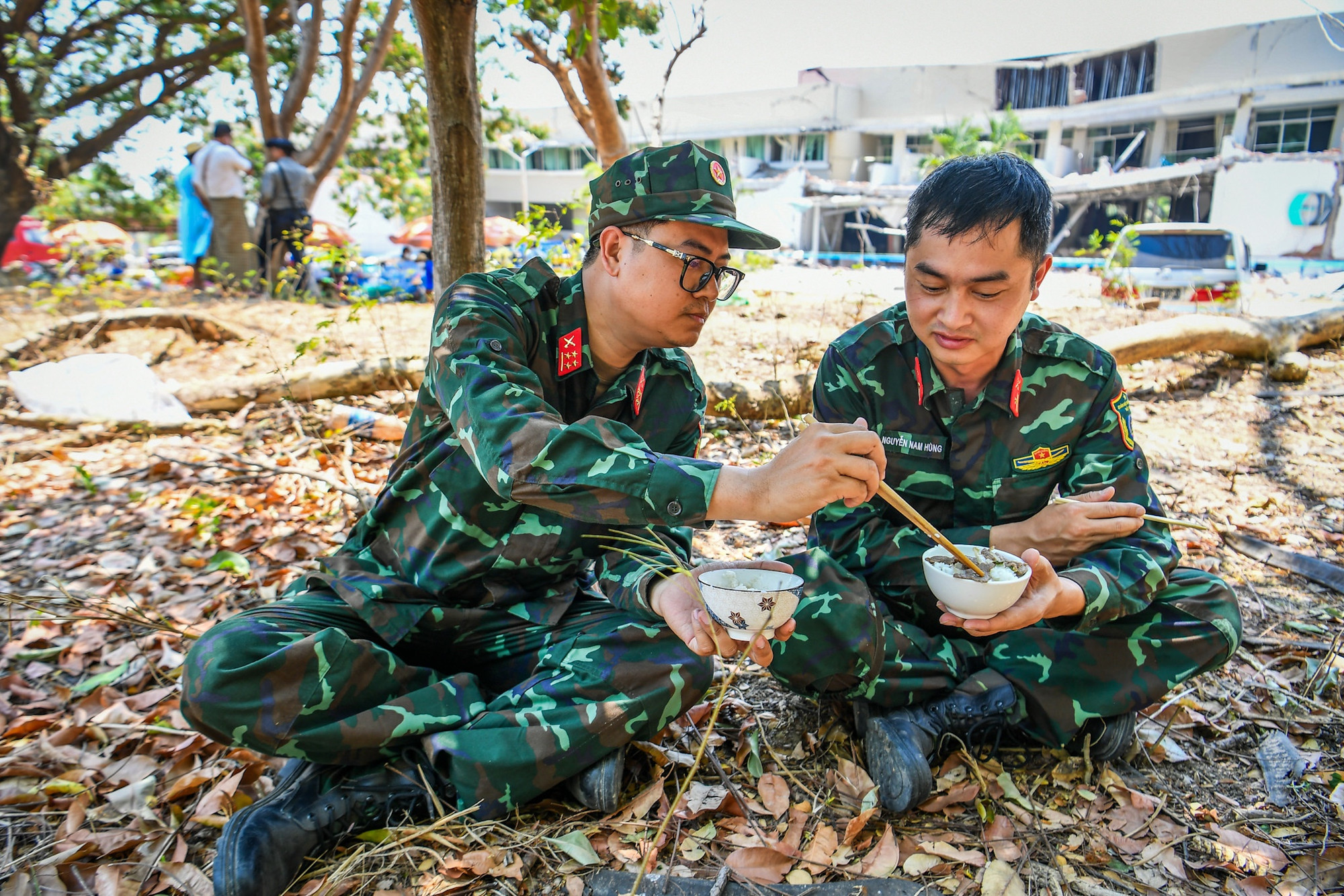 Các cán bộ, chiến sĩ tìm những bóng cây râm mát để dùng bữa. Phía sau là cảnh đổ nát, hoang tàn sau trận động đất ngày 28/3.