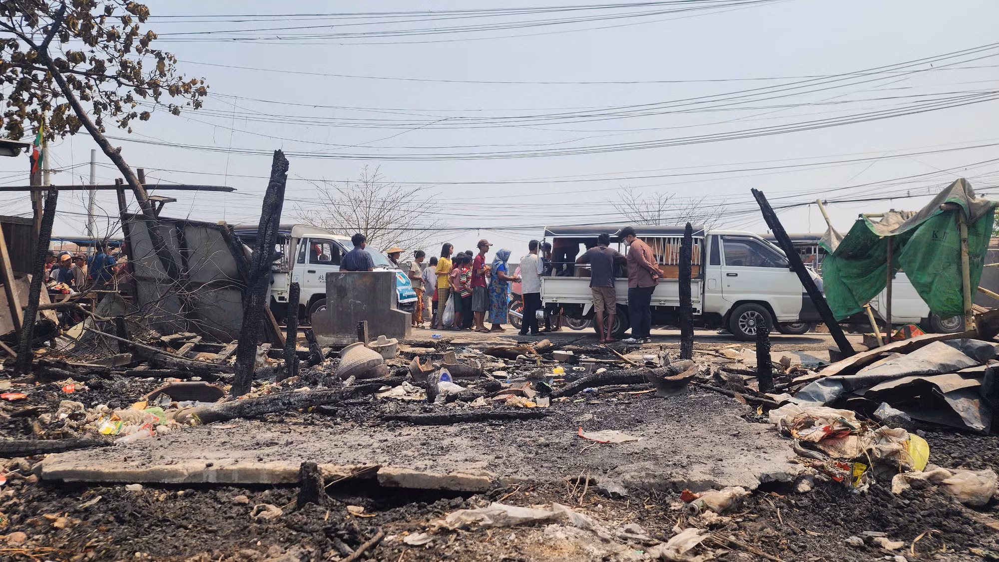 Bên ngoài bãi đất đen đúa vốn là nhà của hàng nghìn người, hai chiếc xe ô-tô đỗ lại để phát đồ ăn trưa, nước uống cho các nạn nhân đã không còn nơi ở... Bên ngoài bãi đất đen đúa vốn là nhà của hàng nghìn người, hai chiếc xe ô-tô đỗ lại để phát đồ ăn trưa, nước uống cho các nạn nhân đã không còn nơi ở...
