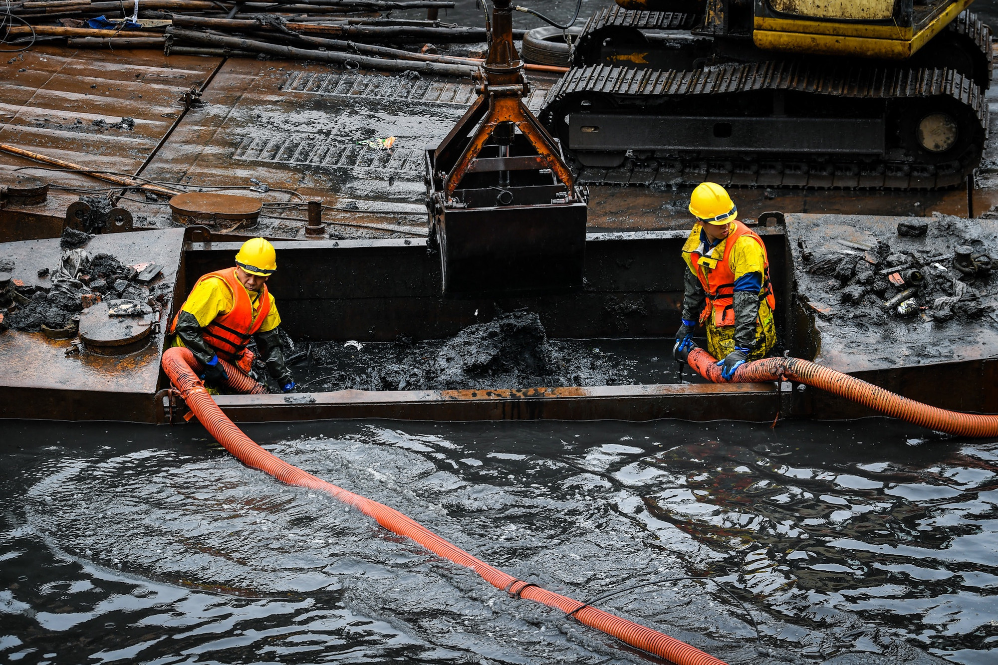 Nửa phần người dưới của các anh ngập sâu trong nước và bùn. Mặc dù vậy, cả hai vẫn cố gắng giữ chặt đầu ống hút để đưa chất thải lên bờ.