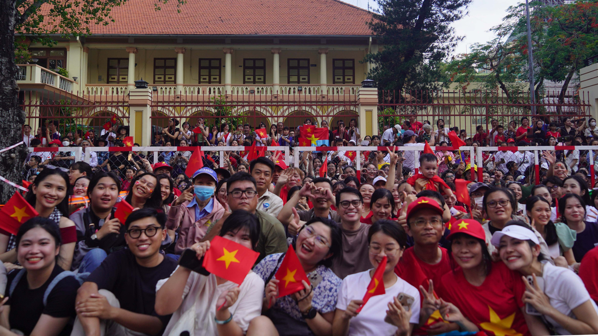 Các em nhỏ theo chân cha mẹ, trên tay cầm cờ đỏ sao vàng, khuôn mặt rạng rỡ chờ đợi khoảnh khắc đoàn diễu binh, diễu hành tiến qua.