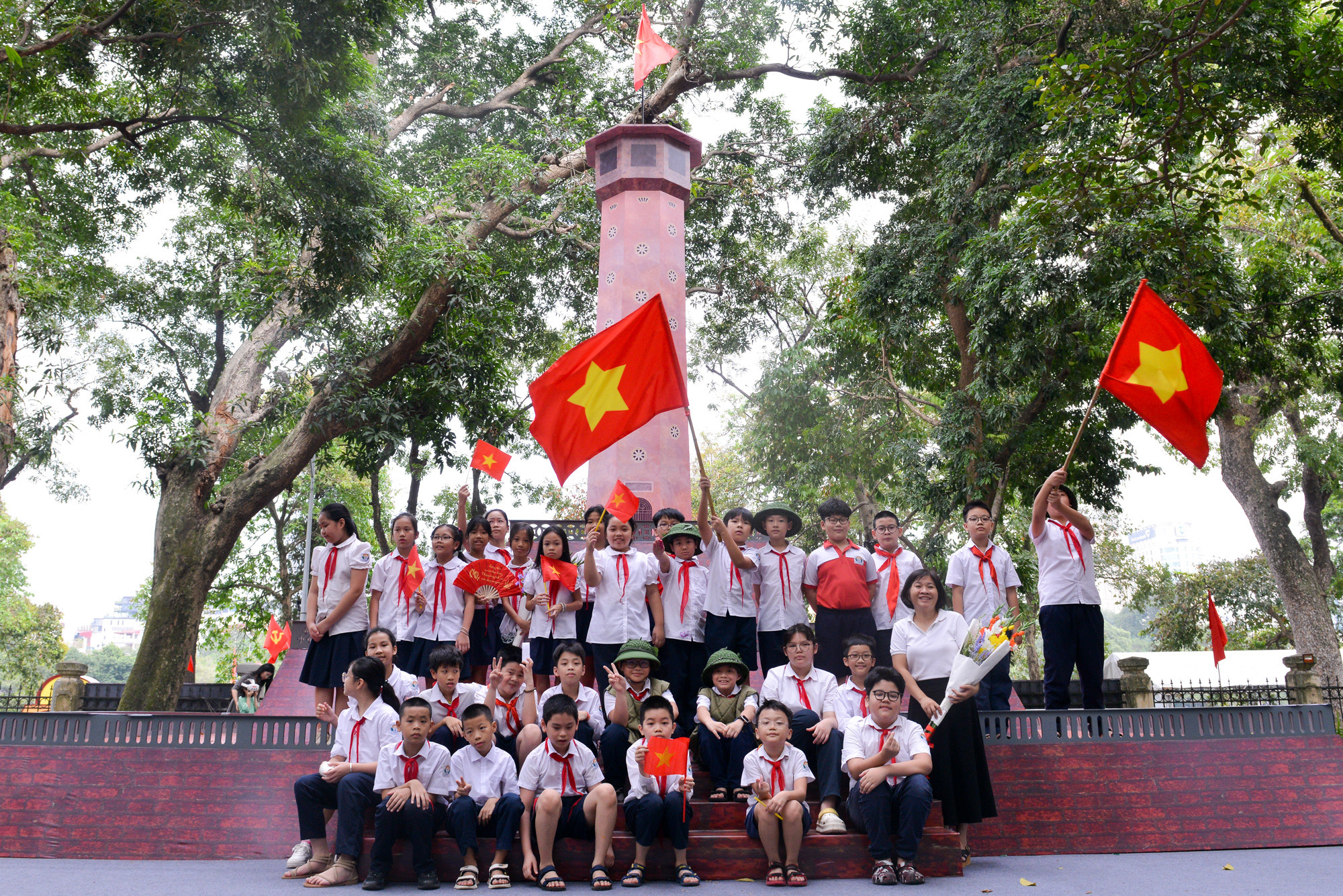 Các bạn học sinh phất cao lá cờ đỏ sao vàng trước mô hình Cột cờ Hà Nội.