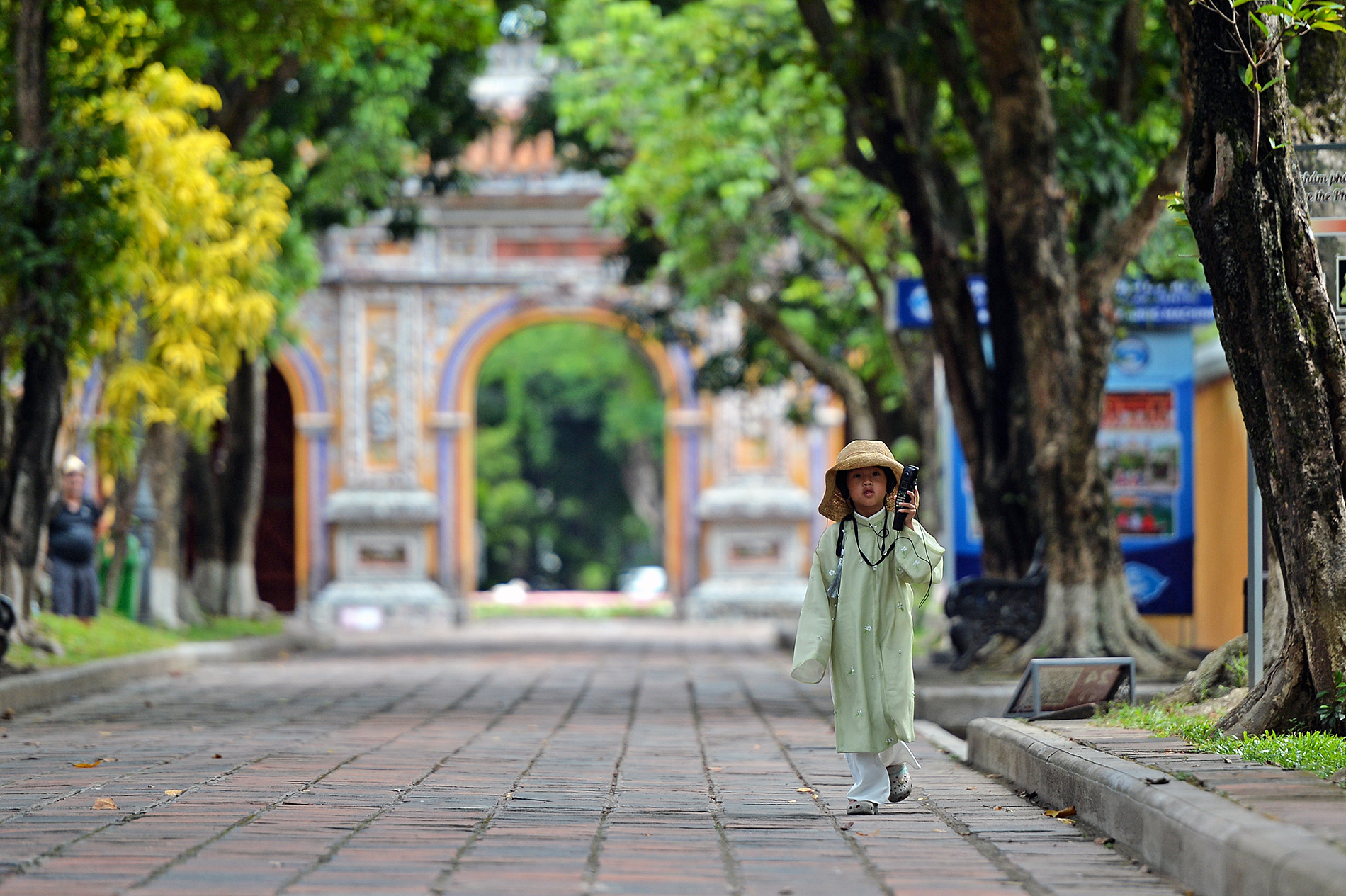 Một cô bé mặc cổ phục lẽo đẽo đi theo mẹ trong khuôn viên Đại nội Huế, trên tai chăm chú lắng nghe lời thuyết trình tự động qua thiết bị phục vụ khách du lịch.