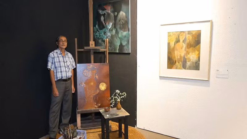 Họa sĩ Hồ Hồng Lĩnh, con trai họa sĩ Hồ Hữu Thủ, bên hai bức tranh dang dở của cha mình được bố trí trong không gian trưng bày . (Ảnh: BTC).