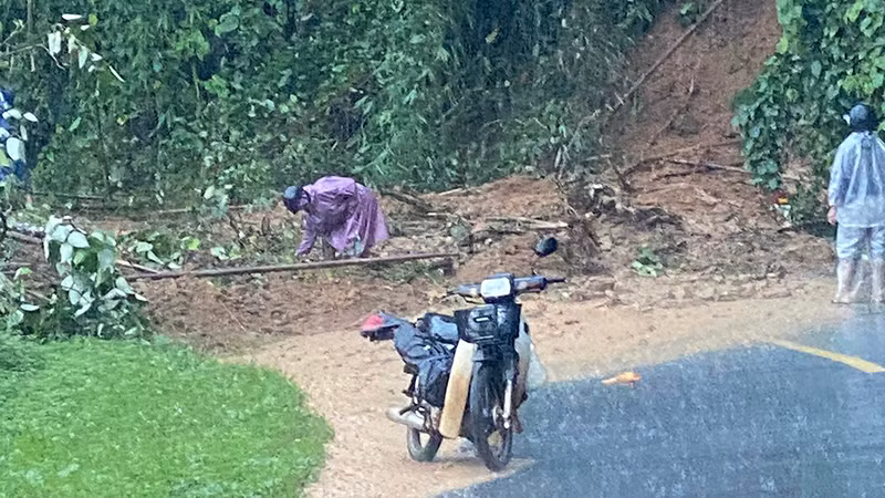 Một số người đi xe máy cố dọn dẹp cây đổ và luồn lách để di chuyển qua khu vực đất đá chắn ngang đường.