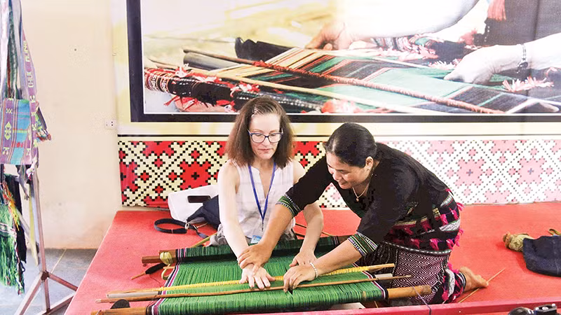 Nghệ nhân H’Bình giới thiệu cách dệt thổ cẩm dân tộc Mạ cho du khách nước ngoài đến tham quan tổ hợp tác.