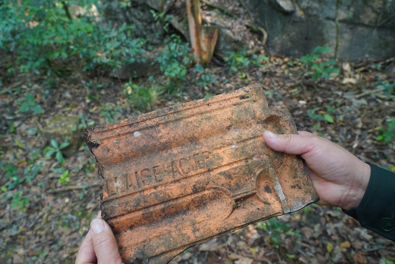 Một viên ngói vỡ của mái lợp hệ thống cáp tời quặng vẫn còn dòng chữ bằng tiếng Pháp sắc nét dù đã nằm lẫn trong cây cỏ hàng trăm năm. (Ảnh: NGỌC TÚ).
