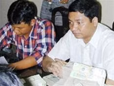 Lê Bảo Quốc (người mặc áo trắng)
ngồi trước đống tiền tang vật,
chờ ký biên bản.