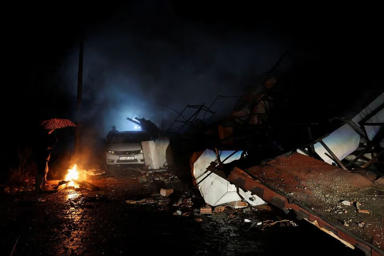 Người dân cầm ô đứng sưởi ấm bên đống lửa tại Antakya, Thổ Nhĩ Kỳ. Điều kiện thời tiết lạnh giá và có mưa đang cản trở lực lượng cứu nạn tìm kiếm các nạn nhân. (Ảnh: Reuters)