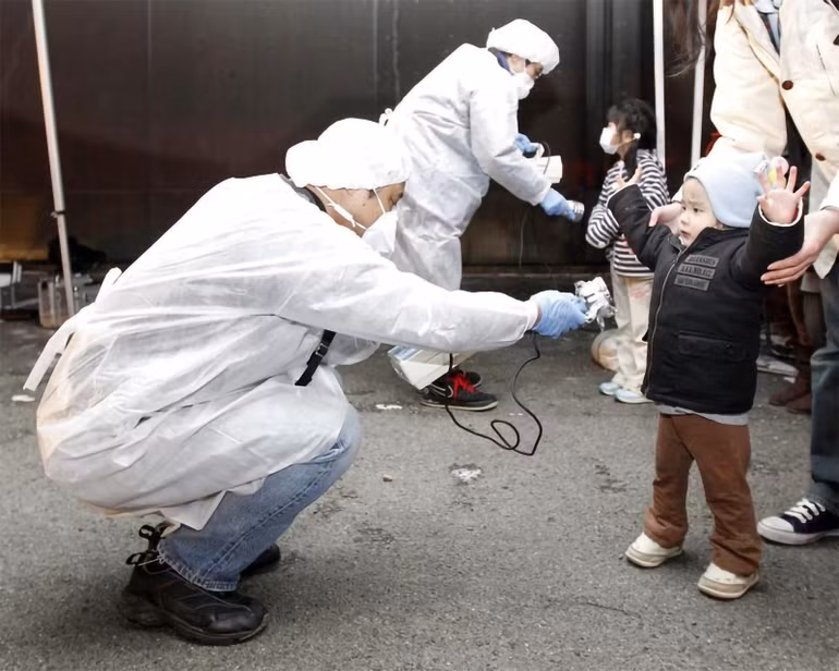 Tại nhà máy điện hạt nhân Fukushima Daiichi ở tỉnh Fukushima, sóng thần đã tràn qua các hệ thống bảo vệ và làm ngập các lò phản ứng, gây ra thảm họa nghiêm trọng. Thảm họa đã làm tê liệt nhà máy điện Fukushima Daiichi, buộc hơn 160.000 cư dân phải sơ tán vì rò rỉ phóng xạ trong không khí. Trong ảnh: Các nhân viên mặc đồ bảo hộ kiểm tra dấu hiệu nhiễm phóng xạ đối với trẻ em đến từ khu vực phải sơ tán gần nhà máy điện hạt nhân Fukushima Daini ở thành phố Koriyama, ngày 13/3/2011. (Ảnh: Reuters)