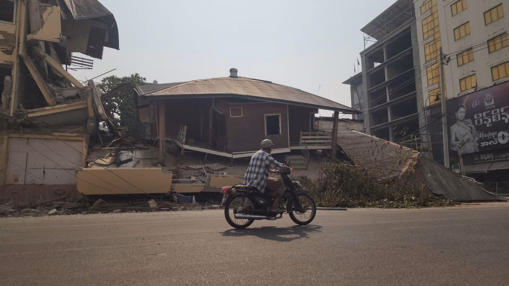 Một người dân địa phương lái xe trên đường phố. Phía sau là những công trình đã không thể ở do ảnh hưởng động đất.